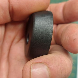 Könnte beinhalten: Ein schwarzes, abgerundetes, zylindrisches Objekt mit einem Loch in der Mitte. Die Oberfläche ist leicht strukturiert. Das Objekt wird in einer Hand gehalten und scheint aus einem festen Material zu bestehen.
