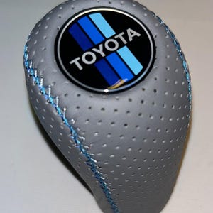 Puede incluir: Pomo de cambio gris perforado con costuras azules. El pomo presenta un emblema circular con el logotipo de Toyota en blanco, azul y negro. El pomo de cambio está diseñado para el cambio de marchas de un vehículo.