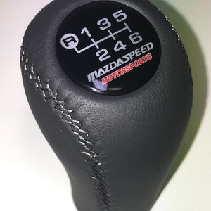 Könnte beinhalten: Schwarzer Schalthebelknauf mit weißem Gangschaltmuster und dem Text "MAZDASPEED MOTORSPORTS" in Weiß und Rot. Der Knauf besteht aus einem dunklen Material mit silbernen Nähten.
