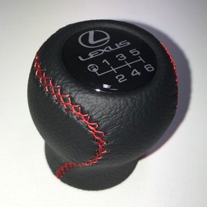 Può includere: Pomello del cambio in pelle nera con cuciture rosse e un logo Lexus nero e argento con schema di cambio marcia.