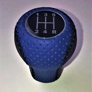 May include: A blue and black gear shift knob. The top of the knob has a black circle with white numbers and lines indicating the gear positions: 1, 2, 3, 4, 5, and R. The knob is covered in a textured blue material.