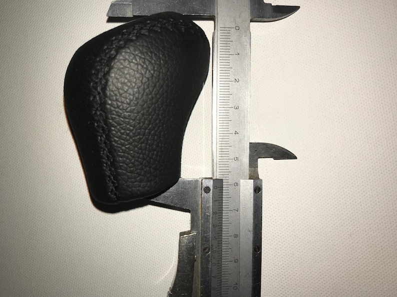 May include: A black leather gear shift knob being measured with a caliper. The caliper reads 6 cm.