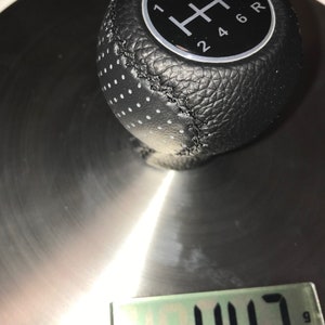 Puede incluir: Una palanca de cambios de cuero negro con un patrón de engranaje plateado y los números 1, 2, 3, 4, 5, 6 y R. La palanca está sobre una báscula plateada que marca 443 gramos.