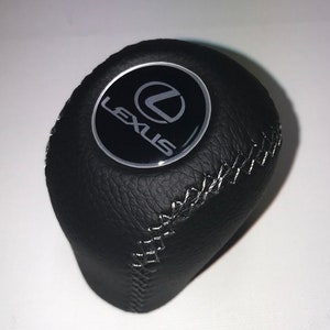 Peut inclure: Pommeau de levier de vitesse en cuir noir avec un logo Lexus argenté et des coutures blanches.