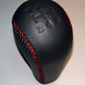 Op de afbeelding: Zwarte versnellingspookknop met rode stiksels. De knop heeft een geperforeerd ontwerp en een reliëfversnellingspatroon: 1, 2, 3, 4, R. Een auto-accessoire.