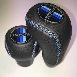 Puede incluir: Dos pomos de palanca de cambios negros con superficie texturizada y costuras cruzadas en azul claro. Cada pomo presenta un emblema circular en la parte superior con la palabra TOYOTA en letras blancas sobre un fondo de franjas verticales azules.