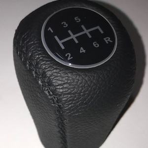 May include: Black leather gear shift knob with a silver and black gear pattern. The pattern shows the numbers 1, 2, 3, 4, 5, 6, and R.