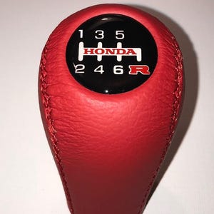 Peut inclure: Un pommeau de levier de vitesse Honda en cuir rouge avec un dessus noir. Le dessus porte des chiffres blancs et le logo Honda. Le schéma de changement de vitesse est 1, 3, 5, 2, 4, 6 et R.