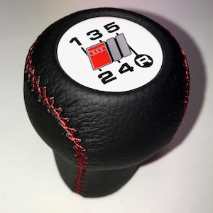 Peut inclure: Pommeau de levier de vitesses en cuir noir avec surpiqûres rouges et un dessus circulaire blanc avec les chiffres 1, 3, 5, 2, 4 et la lettre R. Le dessus comporte également un logo Audi rouge et gris.