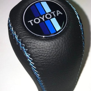 May include: Black leather shift knob with blue stitching. The knob features a circular emblem with the Toyota logo and blue stripes. The design is a sleek, automotive accessory.