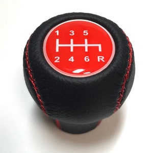May include: Black leather gear shift knob with red stitching. The top has a red circle with a white gear pattern and numbers 1, 3, 5, 2, 4, 6, and R. Designed for a manual transmission.