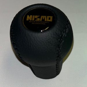 May include: Black leather gear shift knob with a gold NISMO logo and stitching. The logo reads "NISMO Nissan Motorsports International."