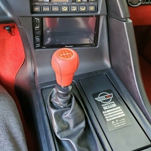 Peut inclure: Un levier de vitesses recouvert de cuir noir avec un soufflet en cuir rouge et un pommeau de vitesse rouge avec un schéma à 5 vitesses. Le levier de vitesses est dans un intérieur de voiture noir avec un autoradio Pioneer et un badge moteur Corvette LT1 avec des spécifications.