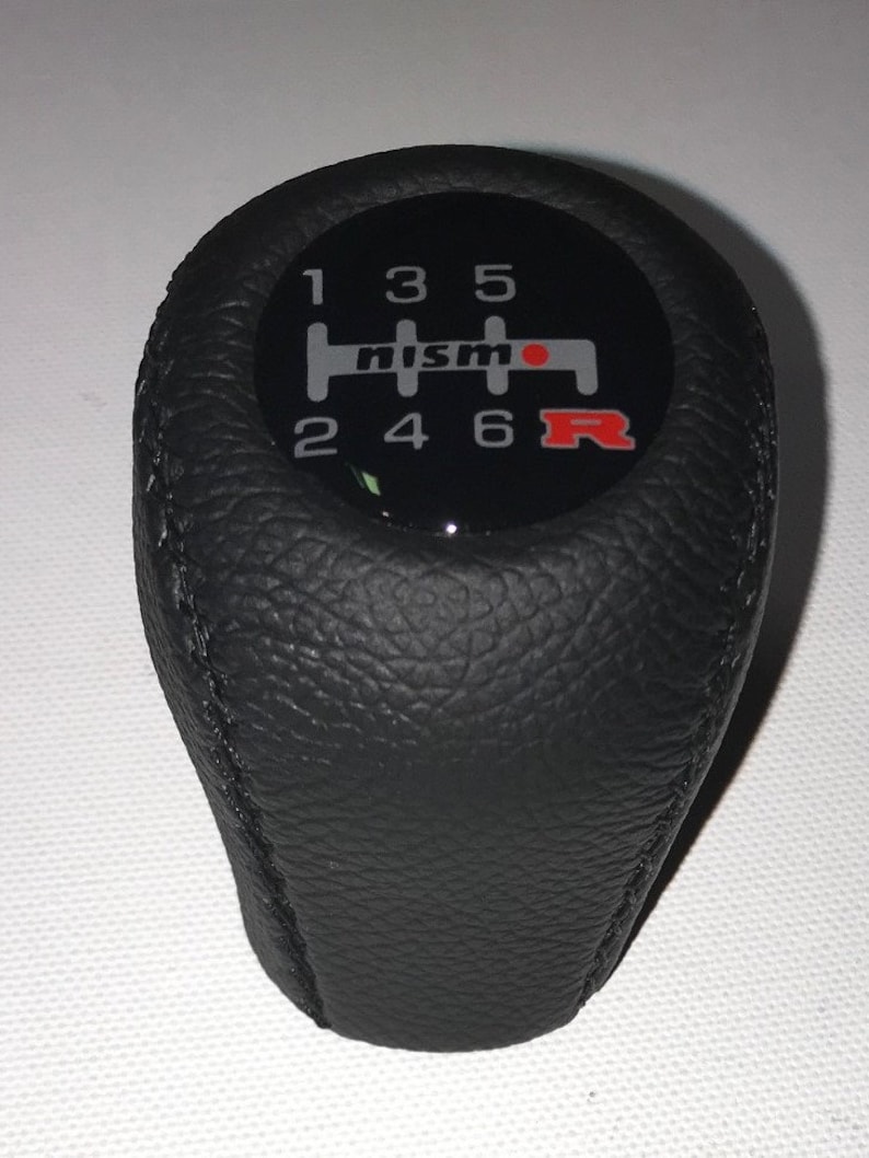 May include: Black leather gear shift knob with a silver and red Nismo logo and a 6-speed gear pattern.