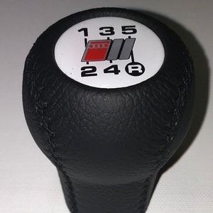 Peut inclure: Pommeau de levier de vitesses en cuir noir avec un dessus circulaire blanc comportant les chiffres 1, 3, 5, 2, 4 et R, ainsi qu'un logo Audi rouge et gris.