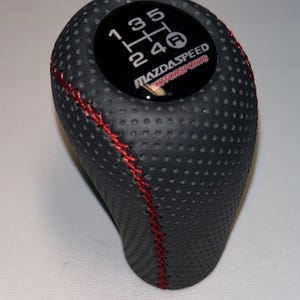 Könnte beinhalten: Schwarzer Schalthebelknauf mit roten Nähten und perforierter Textur. Die Oberseite hat einen schwarzen Kreis mit dem Schaltungsmuster und dem Text "MAZDASPEED MOTORSPORTS".