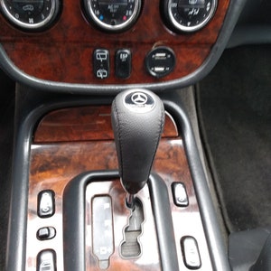 Puede incluir: Primer plano del interior de un coche con un salpicadero de madera y una palanca de cambios de cuero negro. El salpicadero tiene varios botones y diales, incluyendo un panel de control del clima, una radio y una toma de corriente. La palanca de cambios está en la posición 'D'.