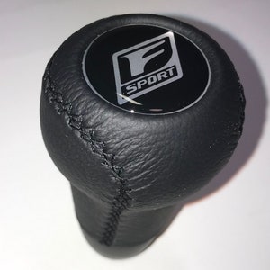 May include: Black leather gear shift knob with a silver F SPORT logo on a black background.