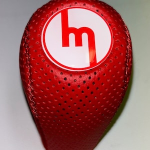 Può includere: Pomello del cambio in pelle rossa con un cerchio bianco e un logo "M" rosso.