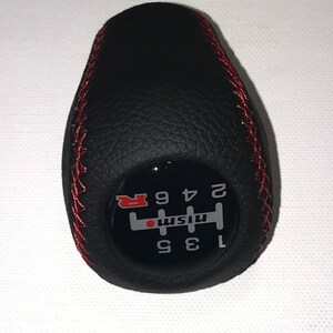 May include: Black leather gear shift knob with red stitching and a black and white gear pattern with the word "Nismo" and the letter "R" in red.