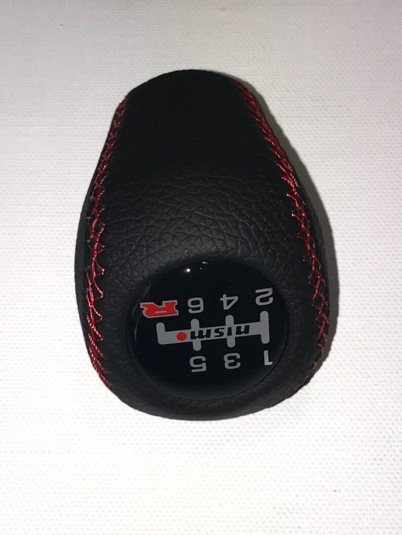 May include: Black leather gear shift knob with red stitching and a black and white gear pattern with the word "Nismo" and the letter "R" in red.