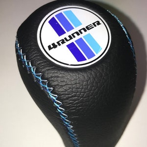 May include: Black leather shift knob with blue stitching. The knob features a white circular emblem with the text "4RUNNER" and blue stripes. The shift knob is designed for a vehicle.