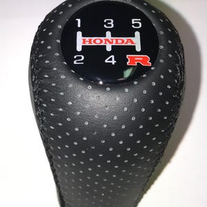 May include: Black perforated leather gear stick knob with a black top displaying the Honda logo in red and white. The gear pattern is shown with white numbers and a red "R".