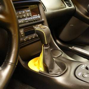 Op de afbeelding: Close-up van een zwart lederen auto-interieur met een gele leren pookknop. De auto heeft een zwart lederen stuurwiel en een zwart dashboard met een radio en klimaatregeling.