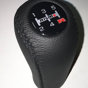 Op de afbeelding: Zwarte leren versnellingspookknop met een zwart-wit Nismo-logo en een rode R op een zwarte achtergrond. De knop heeft witte nummers 1, 2, 3, 4, 5 en R.