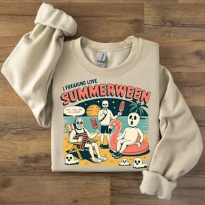 May include: Beige sweatshirt with the text "I freaking love SUMMERWEEN" and a graphic of skeletons on a beach. One skeleton is in a beach chair, another is holding an ice pop, and a third is on a flamingo float.