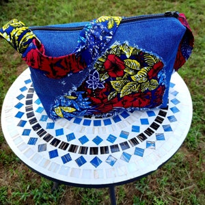May include: A denim handbag with a zipper closure and a colorful floral fabric strap. The bag is sitting on a white and blue mosaic table.