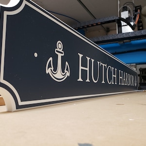 May include: A navy blue sign with white lettering and an anchor design. The sign reads "Hutch Harbor".