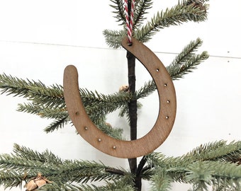 Puede incluir: Un adorno de madera en forma de herradura con un colgador de cuerda roja y blanca. El adorno está colgado de un pequeño árbol de Navidad artificial.