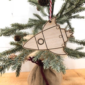 Peut inclure: Une décoration de Noël en forme de fusée en bois avec un cordon de jute rouge et blanc. La décoration est suspendue à un petit sapin de Noël artificiel.