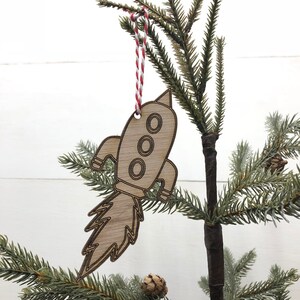 Peut inclure: Ornement de Noël en forme de fusée en bois, suspendu par une ficelle rouge et blanche. La fusée présente trois hublots circulaires et une flamme détaillée. Accroché à une branche de sapin de Noël verte.