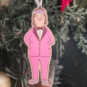 May include: A wooden Christmas ornament featuring a cartoon character in a pink suit, purple bow tie, and a headband. The ornament is hanging from a silver ribbon against a backdrop of a Christmas tree.