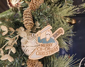 May include: A wooden Christmas ornament featuring a hockey mask-wearing figure holding a machete and baseball bat. The ornament has the text "MERRY CH-CH-CHRISTMAS" and is surrounded by pine needles, pine cones, and decorative gold ornaments.