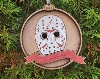 May include: Wooden Christmas ornament featuring a white hockey mask with red accents, a black eye area, and a banner that reads "Merry Ch_Ch Christmas". The ornament is round with a silver ribbon for hanging.