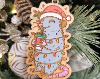 May include: Wooden Christmas ornament featuring a cartoon rat wearing a Santa hat and wrapped in Christmas lights. The rat is holding a coffee cup. The ornament has a silver ribbon for hanging and is set against a Christmas tree background.