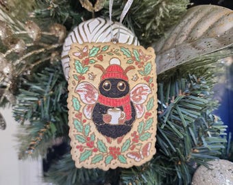 May include: Wooden Christmas ornament featuring a cartoon bee wearing a red hat and scarf, holding a mug. The ornament is decorated with holly and butterflies, and hangs from a silver ribbon on a Christmas tree.