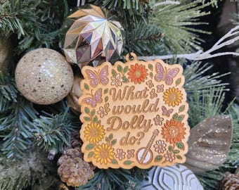 May include: Wooden Christmas ornament with the phrase "What would Dolly do?" surrounded by floral and butterfly designs. The ornament is hanging on a Christmas tree with various ornaments and pine needles.