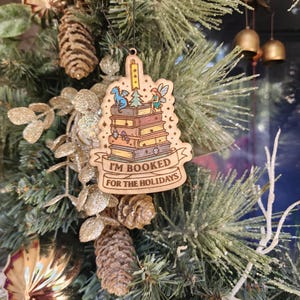 Puede incluir: Adorno navideño de madera con el texto "I'M BOOKED FOR THE HOLIDAYS". El adorno presenta una pila de libros con un dragón, un hada y otros detalles caprichosos. Cuelga de un árbol de Navidad con piñas y adornos dorados.