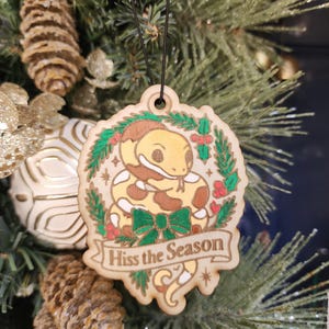Puede incluir: Adorno navideño de madera con un diseño de serpiente de dibujos animados, rodeado de una corona y un lazo. El adorno dice "Hiss the Season" en una pancarta. La serpiente es de color beige y marrón, con una corona verde y bayas rojas. Cuelga de un árbol de Navidad.