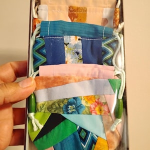 May include: A box containing several patchwork fabric pouches with drawstring fastenings. The pouches display a variety of patterns and colours, including floral, geometric, and solid fabrics. The drawstring cords have wooden beads.