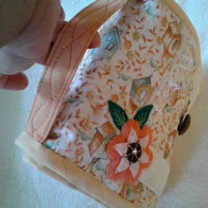 May include: A beige fabric bag with a floral print featuring small houses and birds. The bag has a peach-coloured handle and a white trim. A pink felt flower with a brown button centre is attached to the bag.