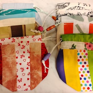 Peut inclure: Plusieurs pochettes à cordon faites à la main, aux couleurs et motifs variés. Les pochettes présentent un mélange de tissus, notamment des rayures, des imprimés floraux et des couleurs unies. Chaque pochette a une fermeture à cordon avec des perles décoratives.