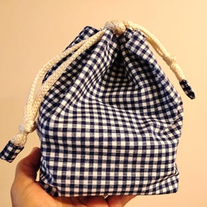 May include: A navy blue and white gingham drawstring bag. The bag features a cream-coloured drawstring closure. The bag is held in a hand.