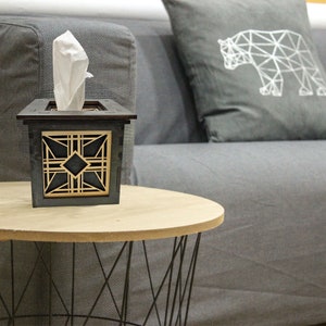 Puede incluir: Un porta pa&ntilde;uelos de madera con un dise&ntilde;o geom&eacute;trico en negro y dorado. El porta pa&ntilde;uelos est&aacute; sobre una mesa de madera con una base de metal negro.