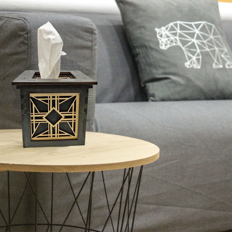 Puede incluir: Un porta pa&ntilde;uelos de madera con un dise&ntilde;o geom&eacute;trico en negro y dorado. El porta pa&ntilde;uelos est&aacute; sobre una mesa de madera con una base de metal negro.
