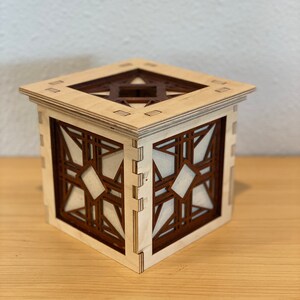 Puede incluir: Una caja de madera con un dise&ntilde;o geom&eacute;trico. La caja est&aacute; hecha de madera clara con detalles en marr&oacute;n oscuro. El dise&ntilde;o es intrincado y presenta una forma de estrella en el centro.